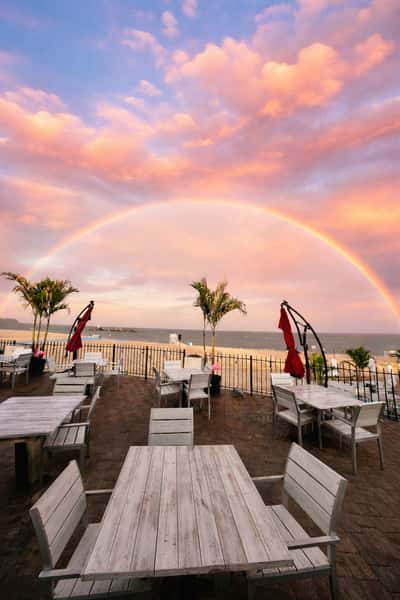 Beachfront patio with white wooden tables, palm trees, and a full rainbow arching across a vibrant sunset sky over the ocean.