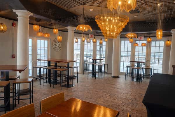 Spacious indoor venue with tall white columns, modern chandeliers, and high-top wooden tables with ocean views through glass doors.