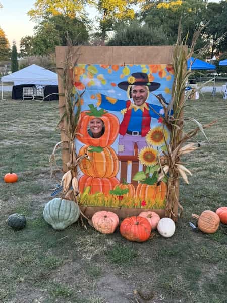 Two people pose with their faces through a festive fall-themed cutout featuring a scarecrow and pumpkins at an outdoor autumn event.