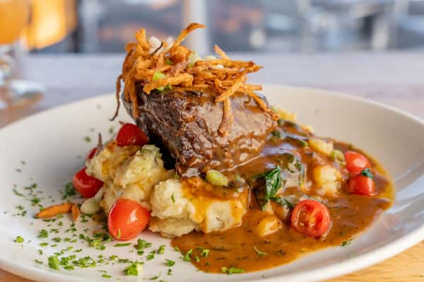 This image shows a beautifully plated dish of braised short ribs served over creamy mashed potatoes, garnished with cherry tomatoes, crispy fried onions, and a savory brown gravy. A sprinkle of fresh herbs completes the presentation.