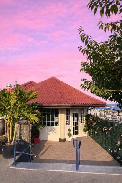 Small stucco building with red tile roof and glass door entrance, set against a vibrant pink sunset sky and tropical landscaping.