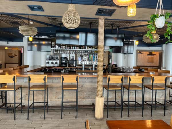 Modern indoor bar with wooden stools, hanging lights, multiple TVs, liquor shelves, and beer taps in a coastal-themed setting.