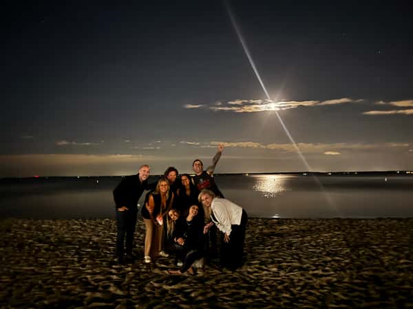 Group of people posing joyfully on a sandy beach at night under a bright full moon reflecting over calm ocean water.