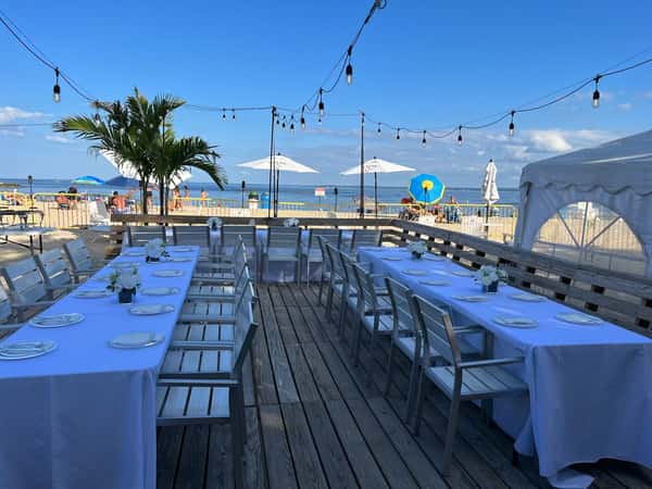 Elegant beachside dining setup with long tables, white tablecloths, string lights, and ocean views under a bright blue sky.