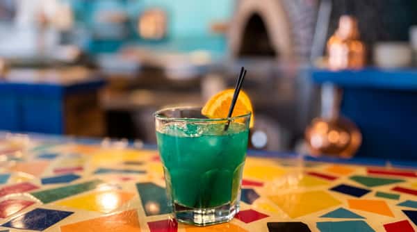 Turquoise cocktail in a glass with ice, an orange slice, and two black straws on a colorful tiled surface.