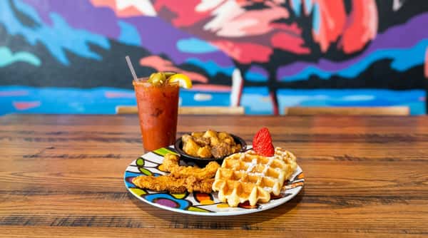 Chicken and waffles with a side of potatoes, and a Bloody Mary with olives and a lemon wedge, on a wooden table.