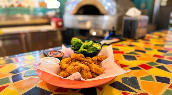 Fried shrimp and a side of broccoli with dipping sauces on a patterned table. A pizza oven is in the background.