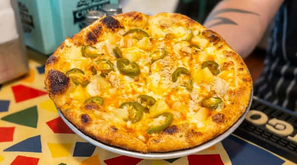 Pizza with pineapple, sliced jalapeños, shredded chicken, and a drizzle of hot sauce on a metal pan.