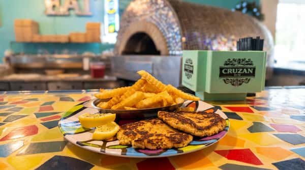 Two crab cakes, crinkle-cut fries, and lemon wedges on a colorful plate, with a mosaic pizza oven in the background.