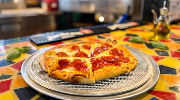 Pepperoni pizza on a metal pan, with a mosaic pizza oven and colorful tabletop blurred in the background.