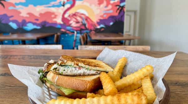 Grilled panini sandwich with creamy filling and lettuce, served with crinkle-cut fries and a blurred flamingo mural in the background.
