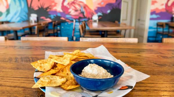 Fried tortilla chips and a blue bowl of white dip on a plate, with a colorful flamingo mural in the background.