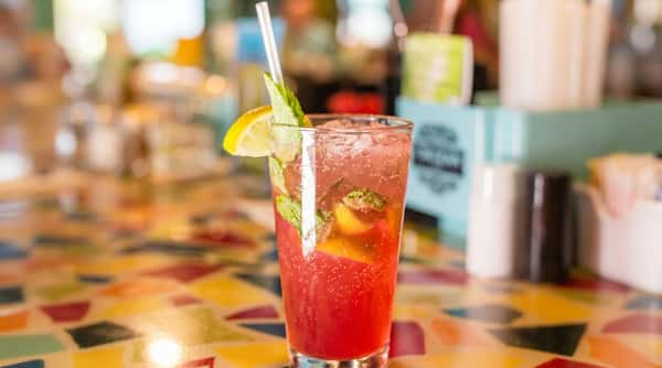 Red cocktail with ice, mint, lemon, and lime slices, with a clear straw, on a colorful patterned table.
