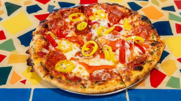 Pizza with pepperoni, red bell peppers, banana pepper rings, and melted cheese on a colorful tiled surface.