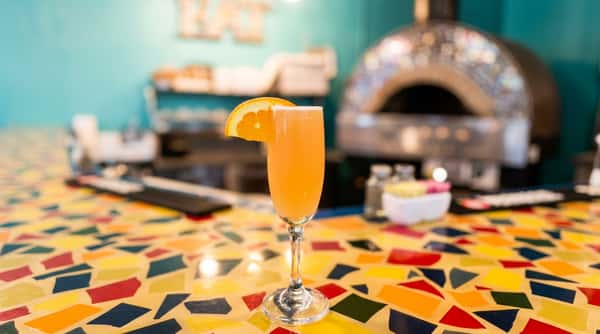 Mimosa cocktail with an orange slice on a colorful mosaic bar top, pizza oven in background.