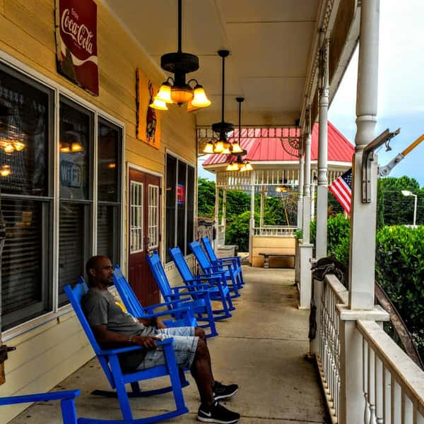 Man relaxing in a bright blue rocking chair on a southern-style barbecue restaurant porch with hanging lights and American flags.