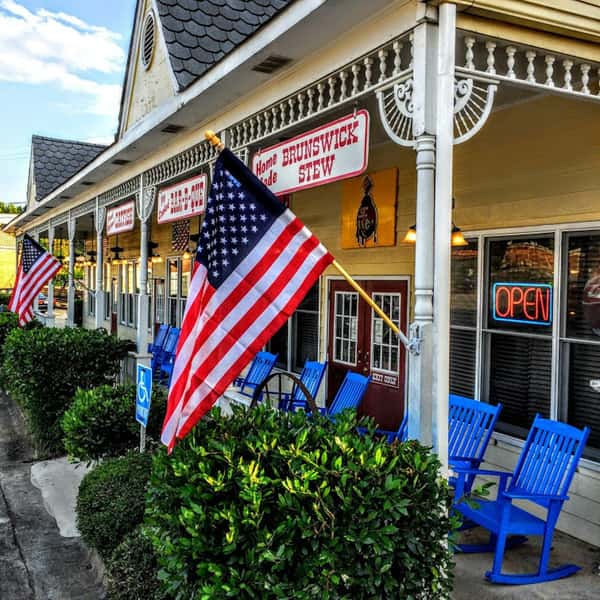 American flags line the porch of a traditional barbecue restaurant with blue rocking chairs and a neon "OPEN" sign.