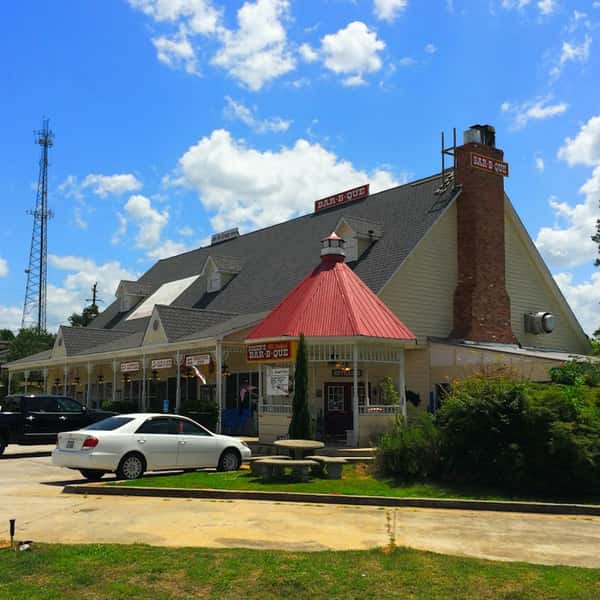 Classic Southern-style barbecue restaurant with a red metal roof, signs reading "BAR-B-QUE," and parked cars out front.