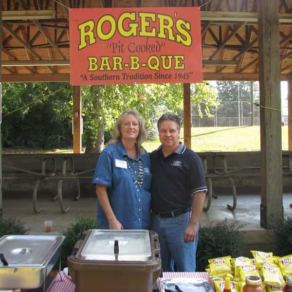 A man and woman stand smiling under a sign for Roger's Bar-B-Que, with picnic tables and food service items in the background.