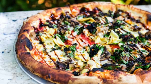 Wood-fired pizza topped with artichokes, mushrooms, greens, and melted cheese on a crisp, charred crust.