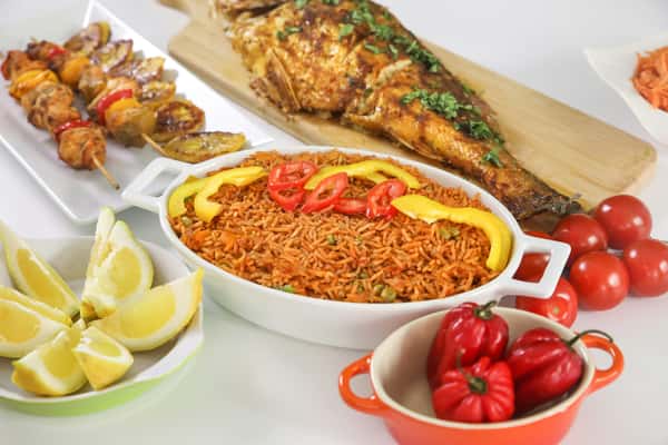 West African party platter featuring jollof rice garnished with red and yellow peppers, grilled whole fish with herbs, chicken and plantain skewers, lemon wedges, and scotch bonnet peppers.