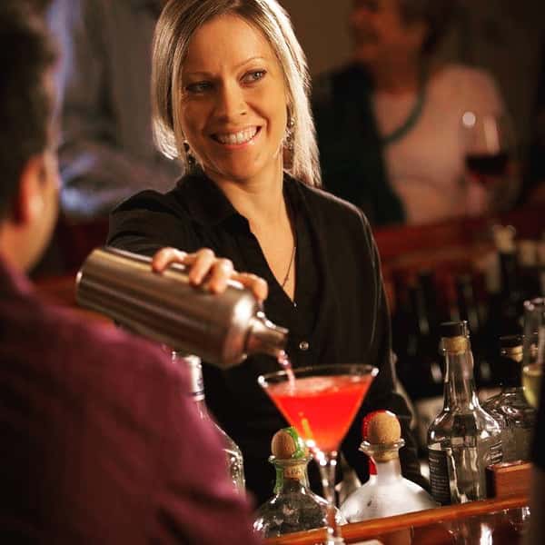 Bartender Faye pouring red cocktail at bar with partial view of several customers