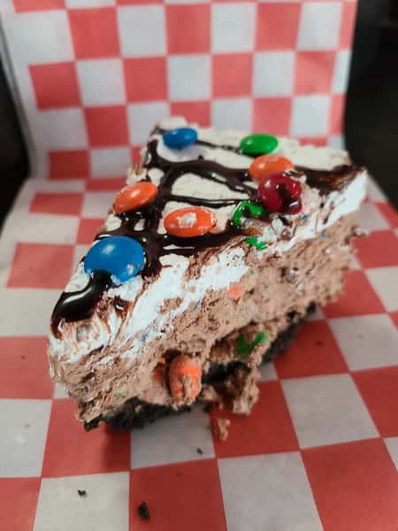 A colorful chocolate pie slice with whipped topping, chocolate drizzle, and multi-colored candies, served on red and white checkered paper.