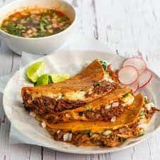 Birria tacos filled with shredded meat and cheese, served with consommé, lime wedges, and sliced radishes on a white plate.