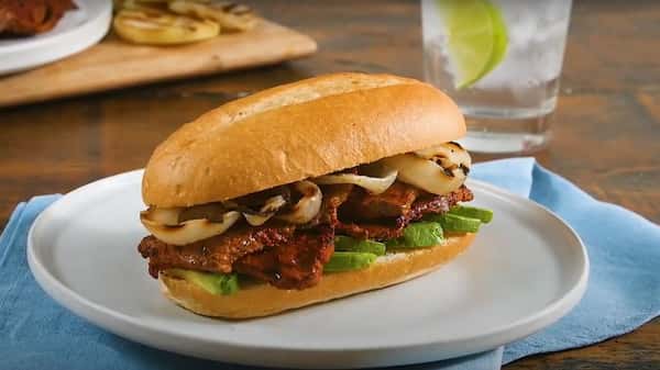 Mexican torta sandwich filled with grilled meat, avocado slices, and sautéed onions on a white plate with a glass of water.
