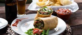 Burrito cut in half served on a plate with lettuce, salsa, and a side of tortilla chips and a drink in the background.