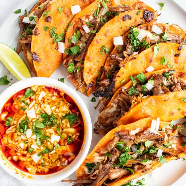 Birria tacos filled with shredded beef, garnished with onions and cilantro, served with a bowl of consommé.