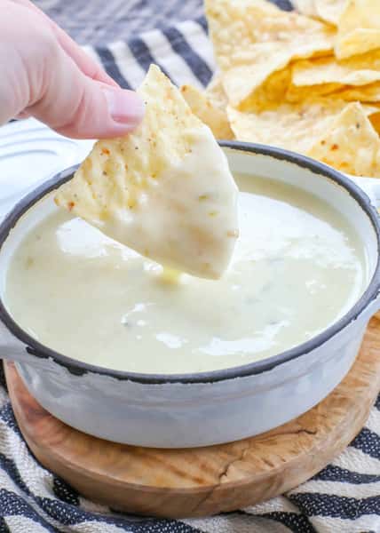 Tortilla chip being dipped into melted white cheese in a metal pot, with more chips in the background.