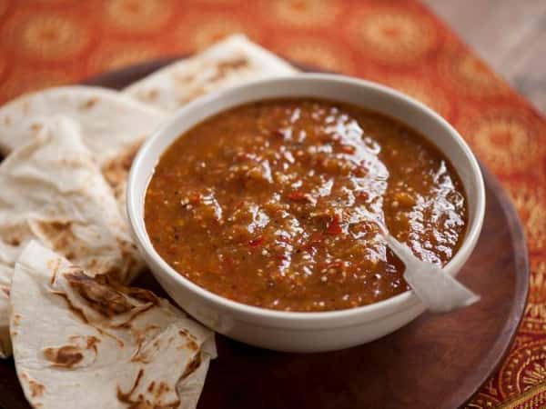 Bowl of roasted tomato salsa with a spoon, accompanied by folded flour tortillas on a wooden platter.