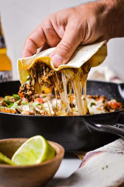Hand lifting a cheesy beef taco with melted cheese stretching from a hot skillet.