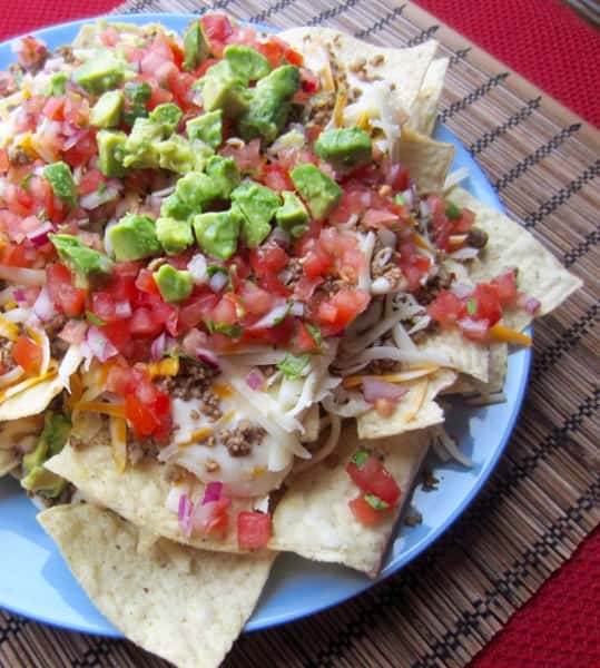Nachos topped with ground beef, cheese, diced tomatoes, onions, and avocado chunks on a blue plate.
