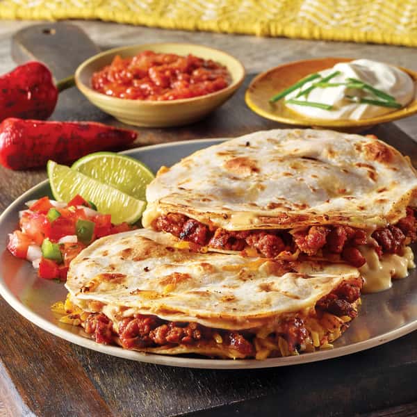 Plate of cheesy beef quesadillas with lime wedges, pico de gallo, sour cream, and salsa in the background.