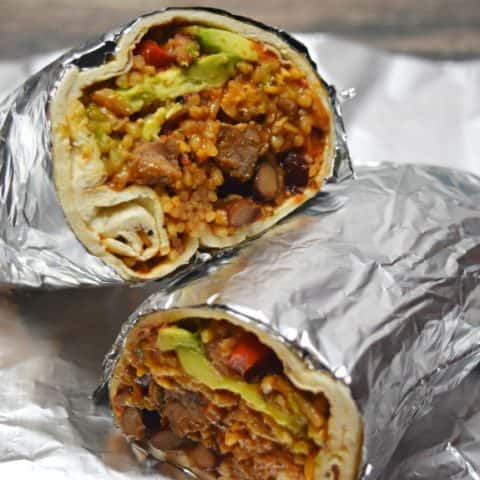 Foil-wrapped burrito cut in half, showing beef, rice, beans, avocado, and vegetables inside.