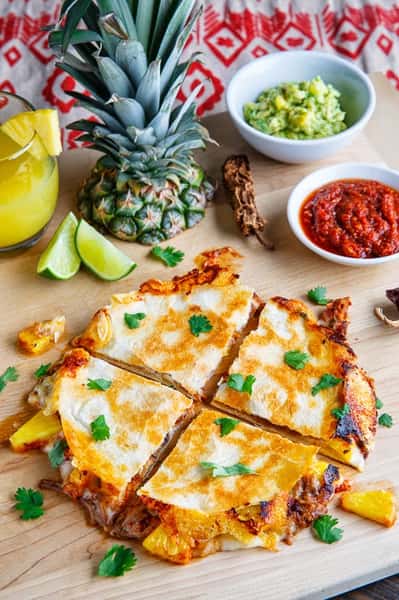 Quesadilla with pineapple and meat garnished with cilantro, served with guacamole, salsa, lime, and a pineapple drink on the side