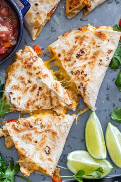 Chicken and cheese quesadillas cut into triangles, served with lime wedges, salsa, and fresh cilantro on a gray tray.