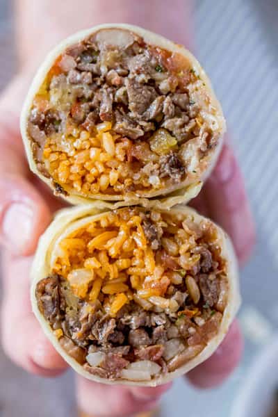 Close-up of a hand holding two burrito halves filled with seasoned rice, grilled beef, onions, and salsa.