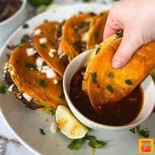 Hand dipping a crispy birria taco into a bowl of consommé.