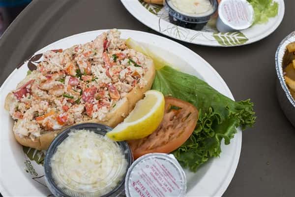 a lobster roll with a side of cole slaw, a lemon wedge, lettuce and a tomato slice