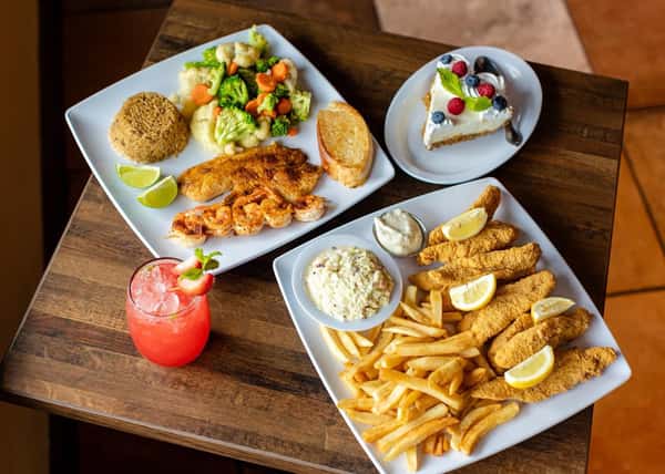 Assorted seafood platters on a wooden table, including fried fish with rice, a dessert, a red drink, and fried seafood with fries