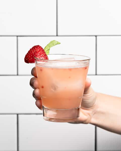 Hand holding a pink cocktail with ice, garnished with a fresh strawberry and mint leaf, against a white tiled background.