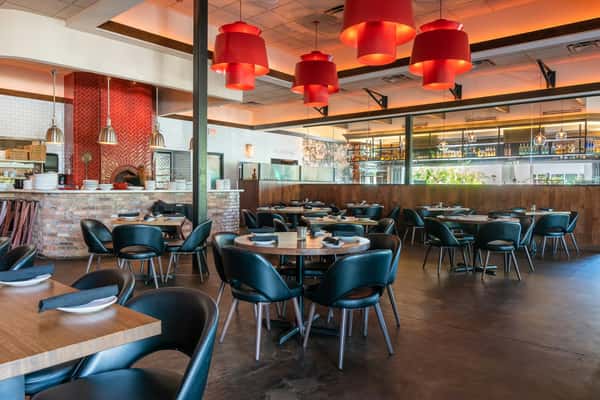 Modern restaurant dining area with black chairs, wood tables, red pendant lights, and a visible brick pizza oven.