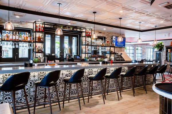 Modern bar with black leather stools, patterned tile front, hanging pendant lights, and shelves stocked with liquor and glassware.