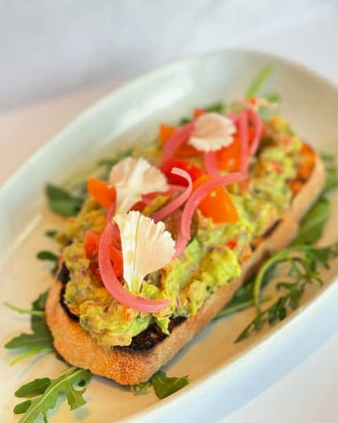 Avocado toast topped with guacamole, pickled onions, diced tomatoes, edible flowers, and served on arugula.