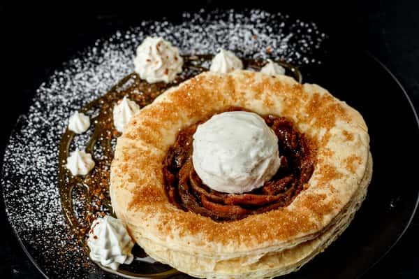 Caramelized apple tart with a puff pastry crust, topped with vanilla ice cream, dollops of whipped cream, caramel drizzle, and powdered sugar.