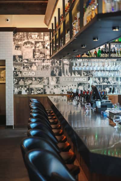 Modern bar interior with black leather stools, a polished counter, hanging glassware, and a wall of vintage black-and-white photos.