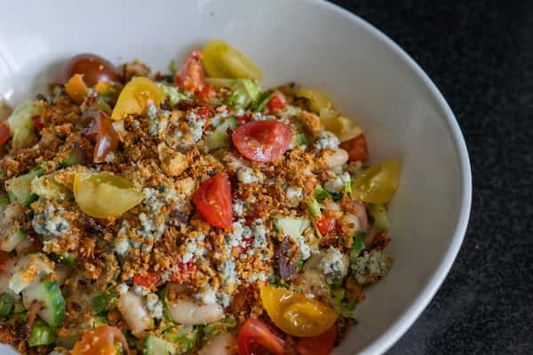 Chopped salad with cherry tomatoes, cucumbers, white beans, blue cheese crumbles, and crispy topping in a white bowl.
